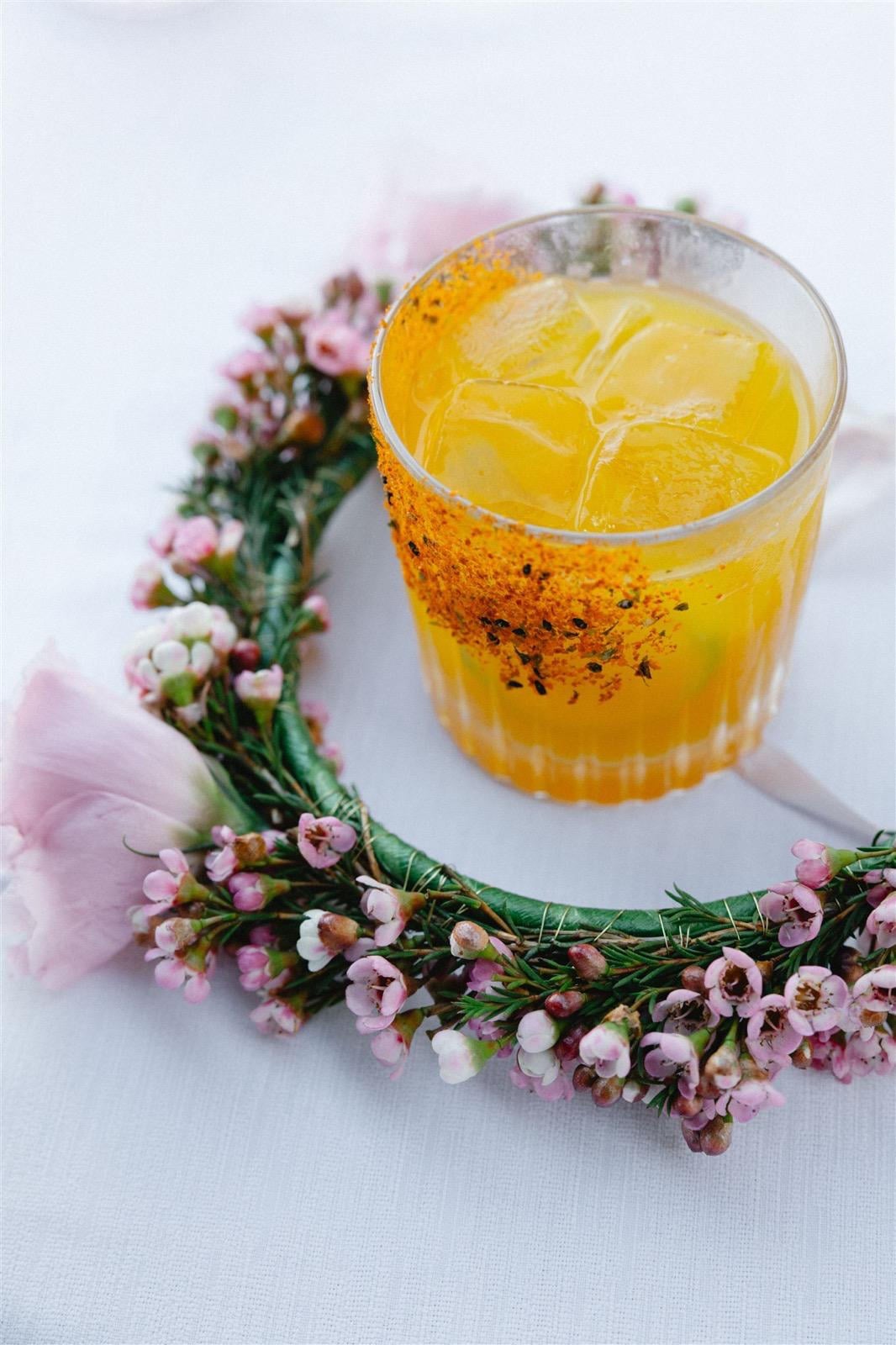 A yellow cocktail garnished with spices and surrounded by a floral wreath